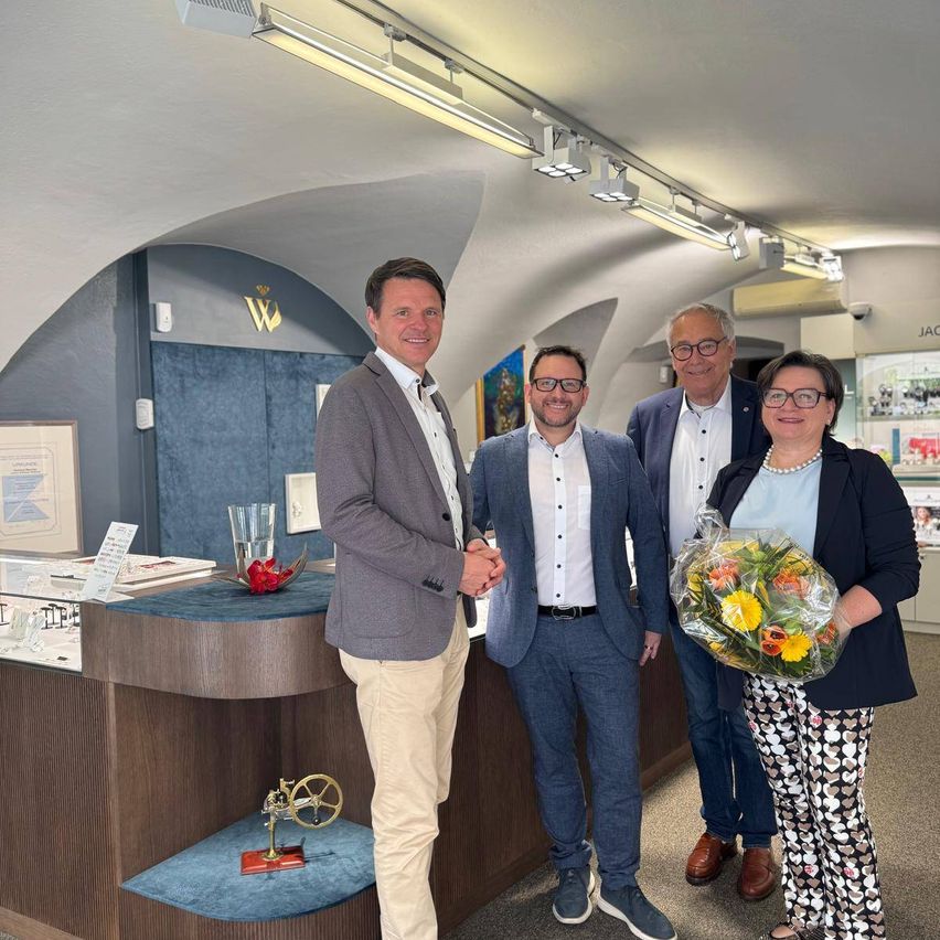 Four men and a woman are standing in a room, smiling and posing for a photo. They are wearing formal attire, and a woman is holding a bouquet of flowers. Behind them, a reception desk is placed, and a few trophies are placed on it. A lamp is mounted on the ceiling.