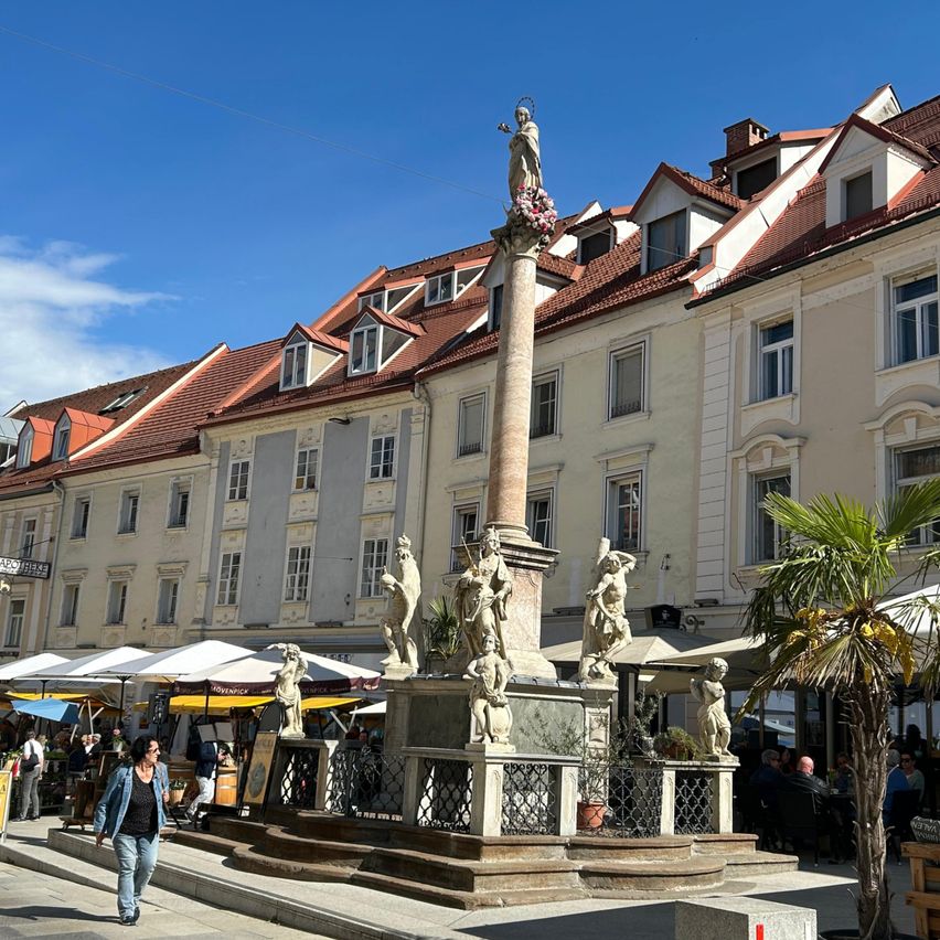 A statue of a woman is atop a column in a town square. There are sculptures around the base. People walk past while others sit in a cafe. Buildings with red roofs and white trim surround the square.
