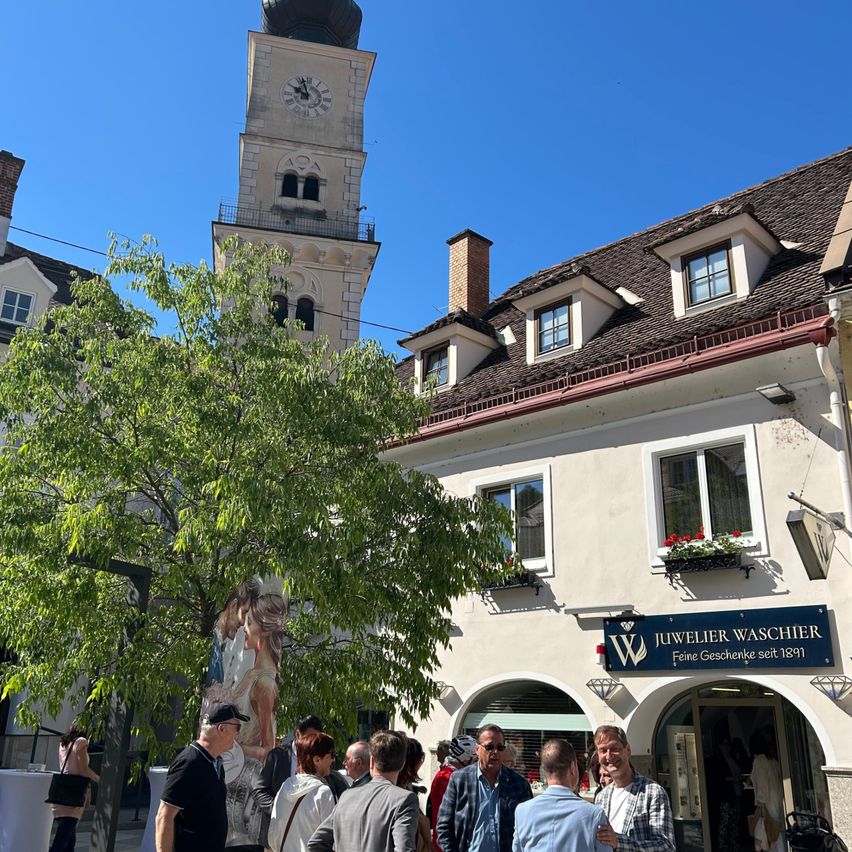 A group of people gather outside a building with a sign 'Juwelier Waschier'. Behind them, there is a tall clock tower, and a tree stands in front of the building.