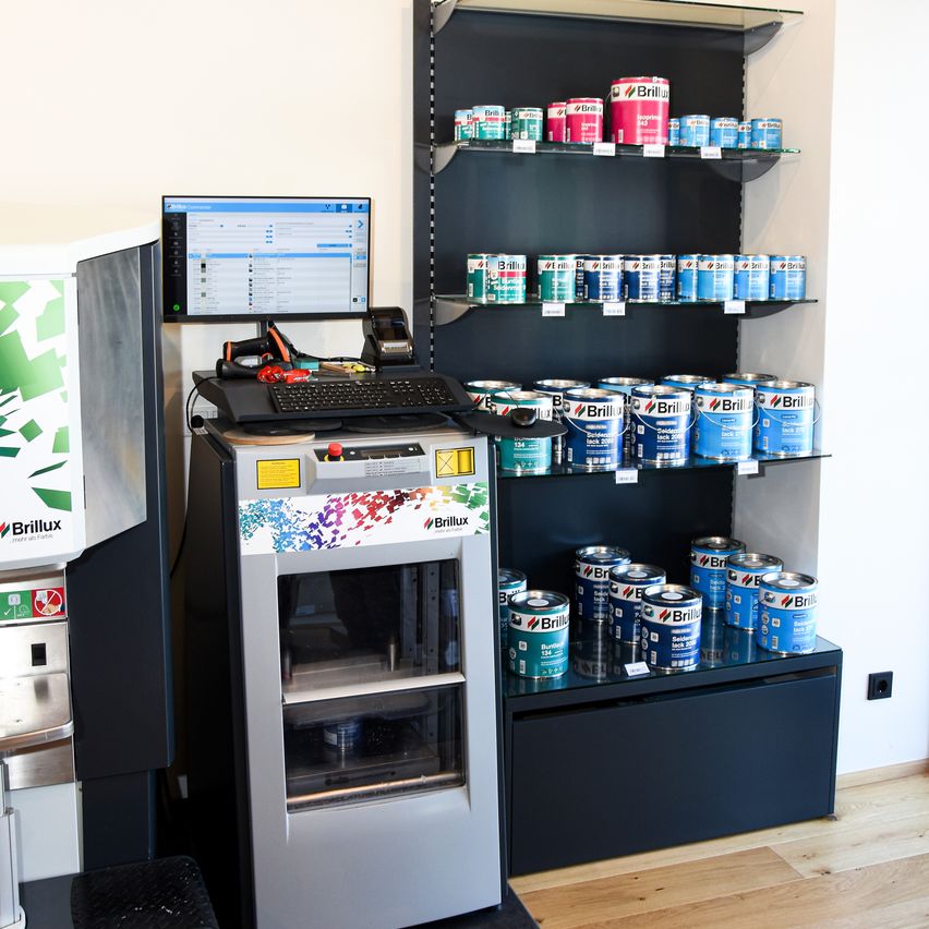 A machine with a computer monitor and a keyboard stands next to a shelf filled with cans of paint. The paint cans are arranged in rows on the shelves.
