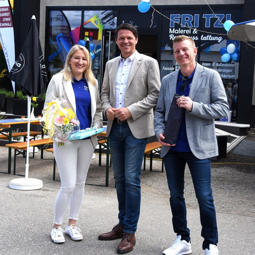 Three people stand together outside a building with a sign reading 'FRITZI Malerei & Raumgestaltung'. A woman holds a bouquet of flowers, while two men smile at the camera. Behind them, tables and chairs are set up with balloons and flags.