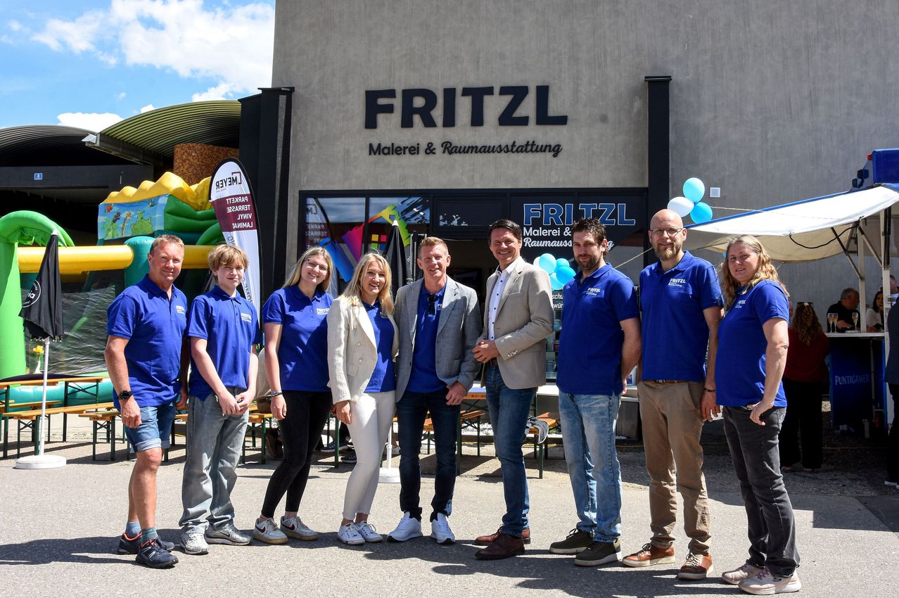 A group of people, including men and women, are standing outside a Fritzl store. They are wearing blue shirts and pants. Behind them, there is a banner, a balloon, and an inflatable play area.