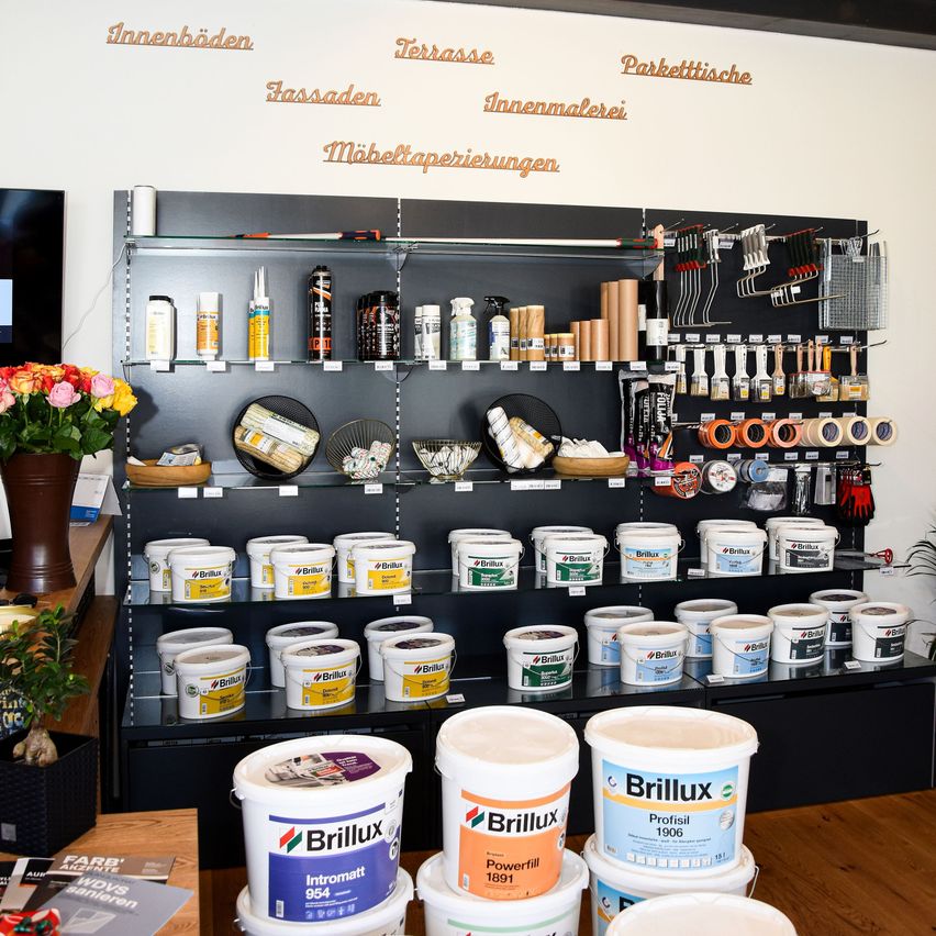 A store displays a variety of paint buckets, labeled with brands and colors. Shelves hold tools, and a bouquet of flowers adds a decorative touch. Words on the wall indicate different areas of application.