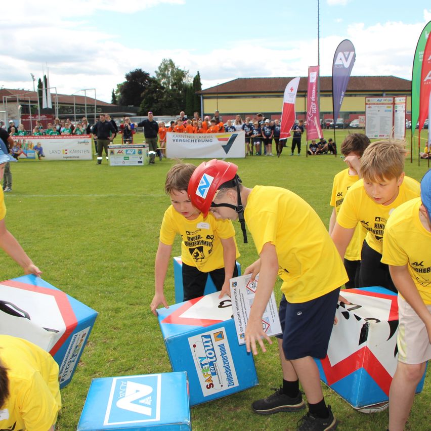 A group of children in yellow shirts are playing a game on a field with large boxes. One child is wearing a red helmet and is leaning over a box. There are more children and adults in the background.