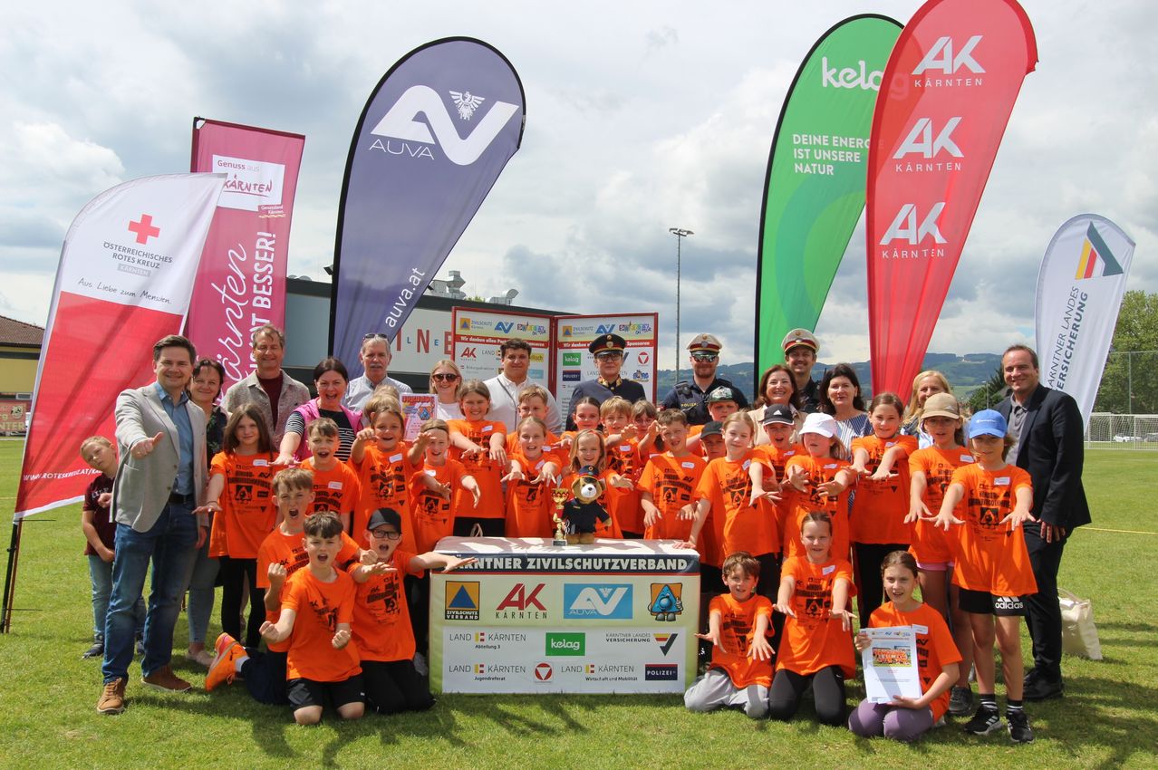 A group of children and adults in orange shirts pose for a photo with a banner in the background. A man holds a trophy, while others wear hats and sunglasses.