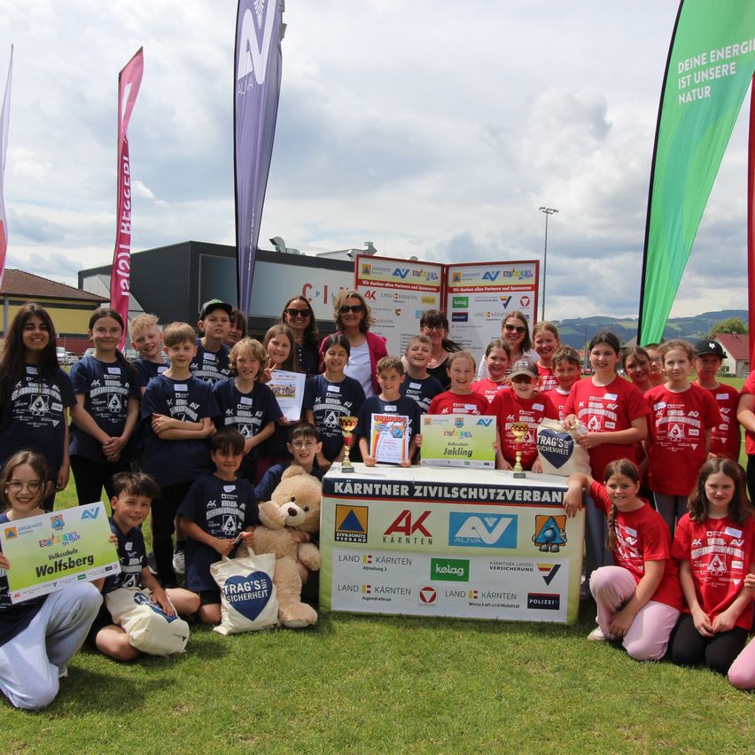 A group of children and adults posing for a photo with a table and prizes. The children wear red and blue shirts. A teddy bear is in front of the table.