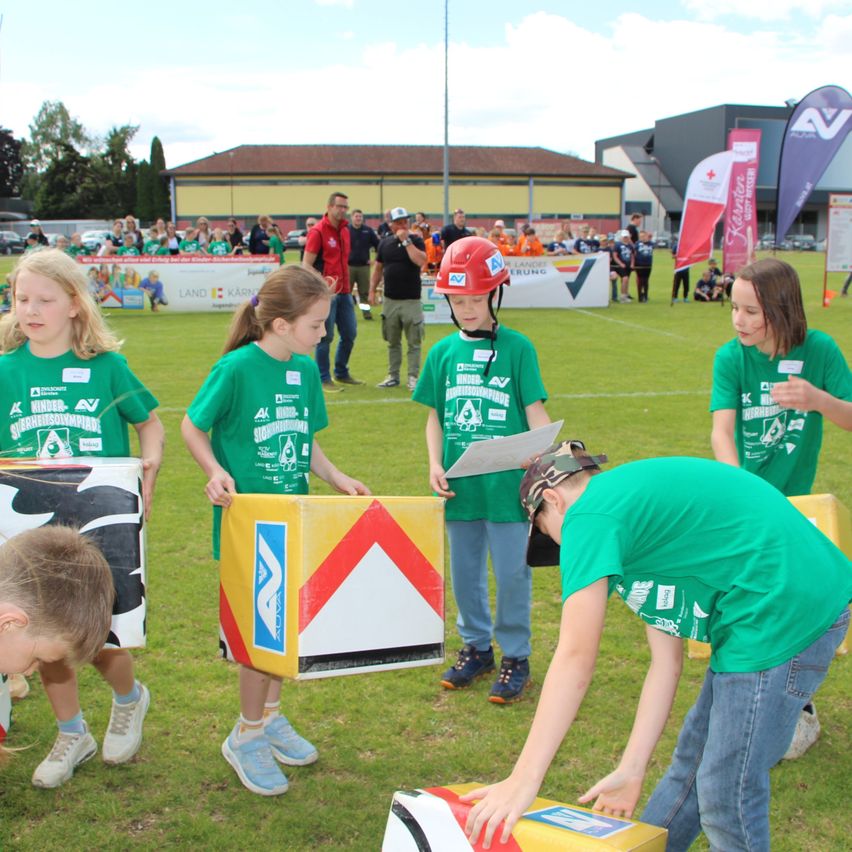 Kids in green shirts and helmets participate in an outdoor activity on a field, with a man in a green shirt checking something on a yellow and red box.