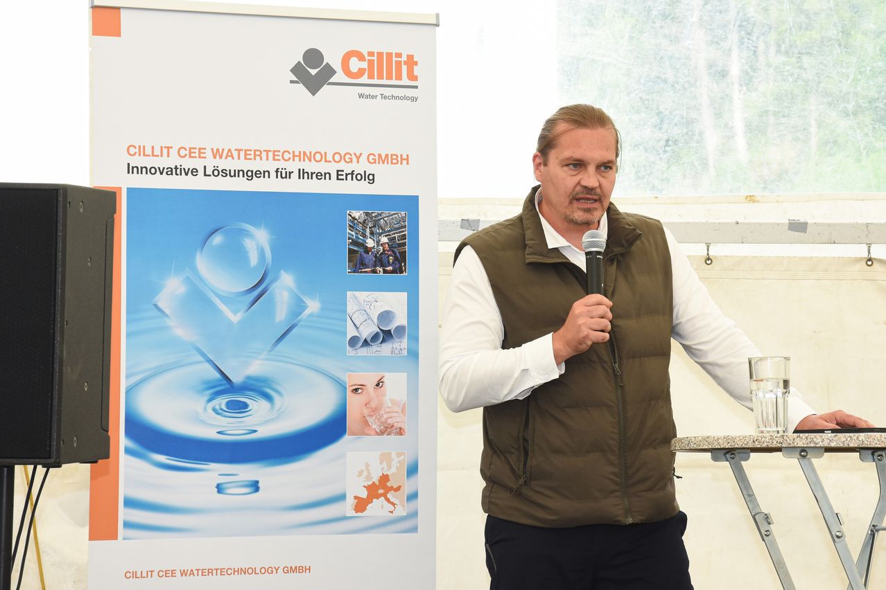 Ein Mann in einer braunen Weste steht an einem Rednerpult, hält ein Mikrofon in der Hand, vor einem Banner, das Cilllit CEE Watertechnology GmbH bewirbt.