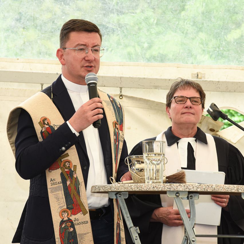 Ein Priester spricht in ein Mikrofon, während ein anderer ein Buch in der Hand hält. Beide tragen Klerikerroben. Hinter ihnen steht ein Tisch mit einem Glas und einem Buch.