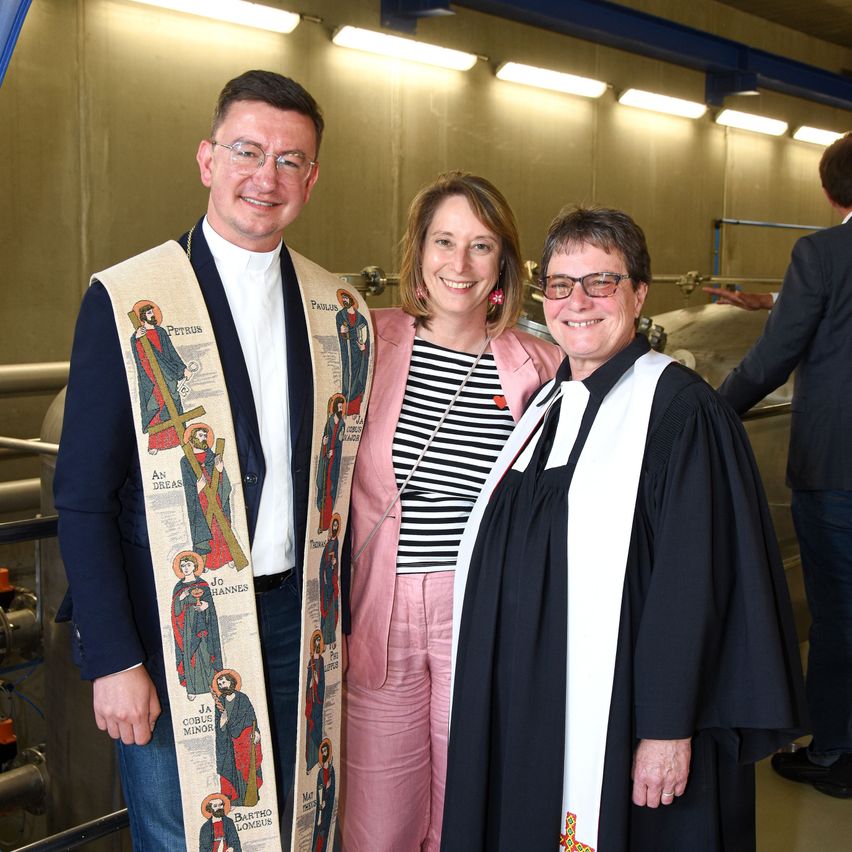 Drei Personen stehen in einem Raum mit metallischem Hintergrund. Zwei von ihnen tragen klerikale Kleidung. Der Mann hat eine weiße Stola. Die Frau trägt einen rosa Blazer und ein schwarz-weiß gestreiftes Hemd.