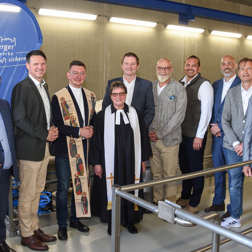Eine Gruppe von Männern, einschließlich eines Priesters, posiert für ein Foto in einer industriellen Umgebung mit einem Banner und Lichtern im Hintergrund.