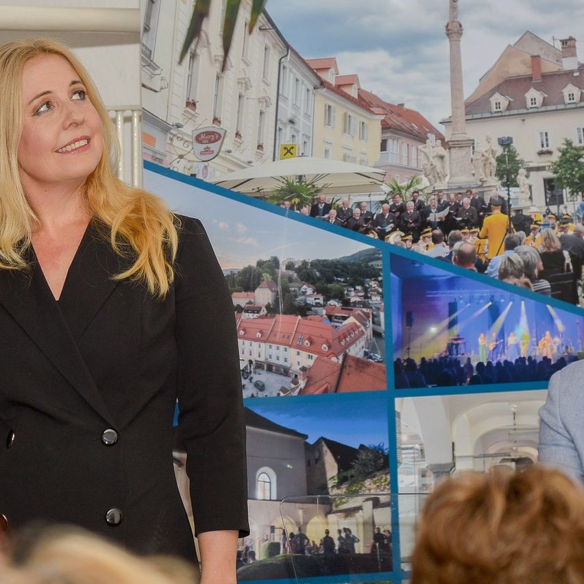 Eine Frau in einem schwarzen Blazer spricht vor einer großen Collage von Bildern, die eine Stadt mit Menschen und Gebäuden darstellen.