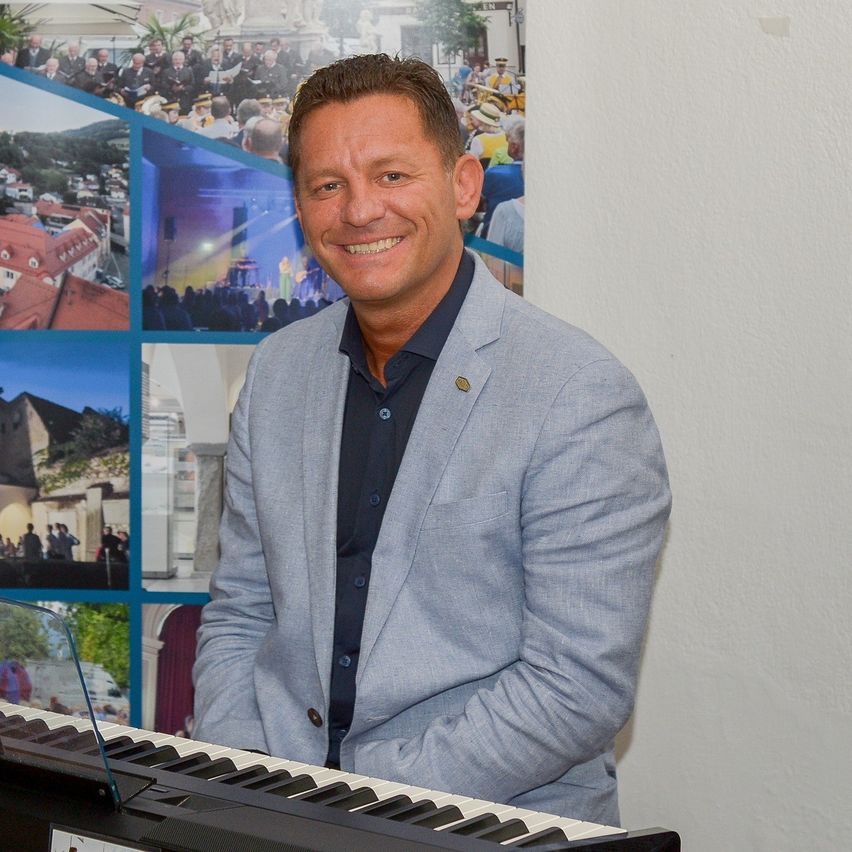 Ein Mann in einem blauen Anzug steht lächelnd vor einem Klavier, mit einer Kulisse aus verschiedenen Veranstaltungsfotos.