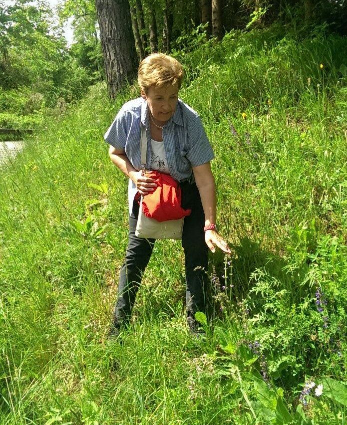 Eine ältere Frau pflückt Blumen in einem Grasfeld, trägt eine rote Tasche und eine Halskette.