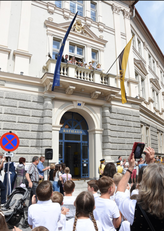Menschenmenge vor dem Rathaus, mit Fahnen und Balkonen sichtbar, eine Gruppe macht Fotos.