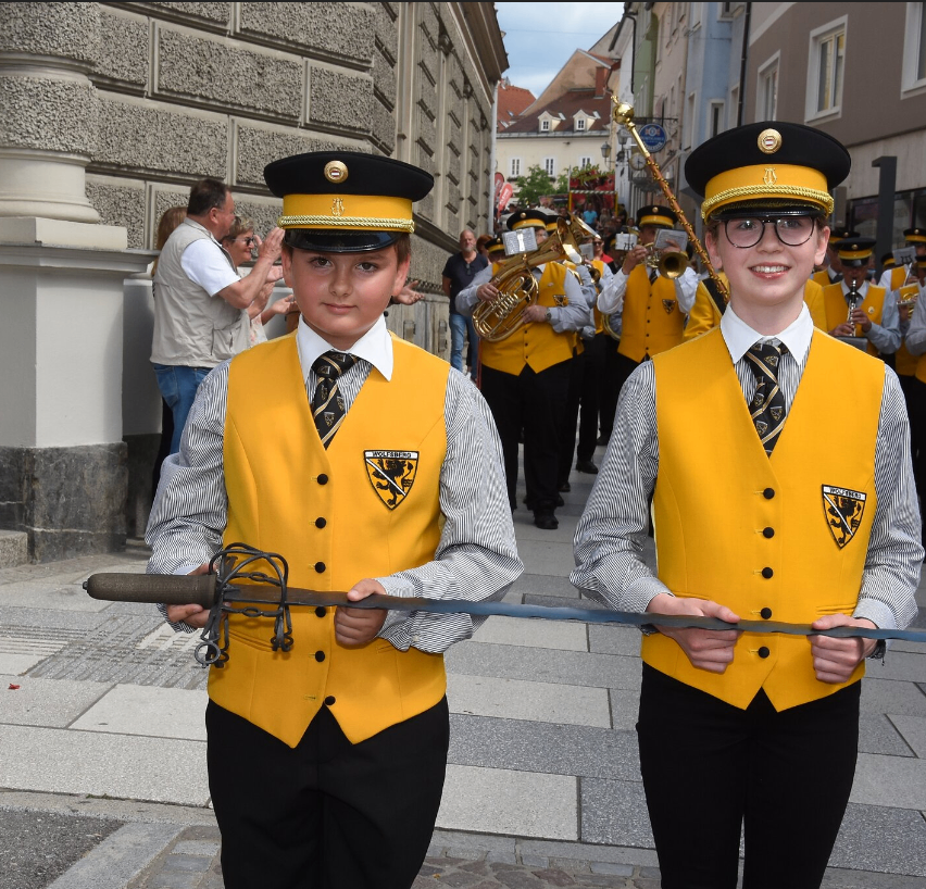 Zwei Jungen in gelben Westen, Schwerter in der Hand, stehen vor einer Marschkapelle auf der Straße. Hinter ihnen beobachtet eine Menge, während Musiker Instrumente spielen.