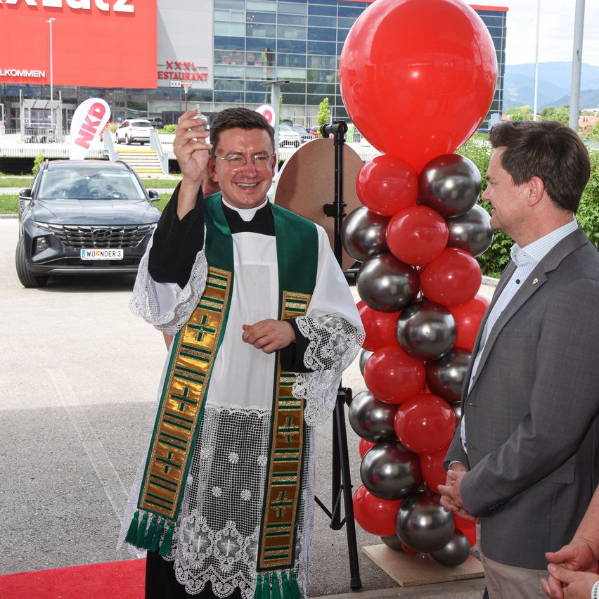 Ein Priester und ein Mann im Anzug stehen draußen in der Nähe eines Autos und Ballons. Hinter ihnen steht ein Gebäude mit einem Schild und einem geparkten Auto.