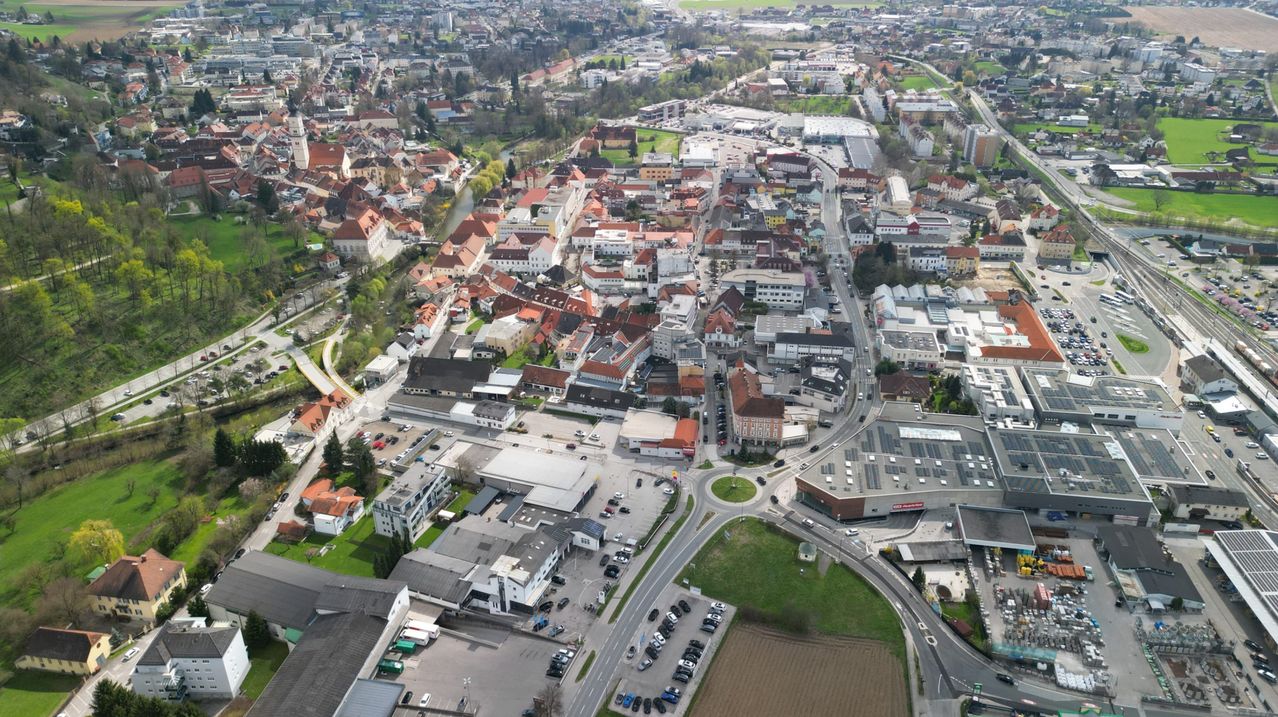 Aerial view of a city with many buildings, some with red roofs, and a river flowing through it. The city is surrounded by trees and grass. Cars are parked in the parking lots.