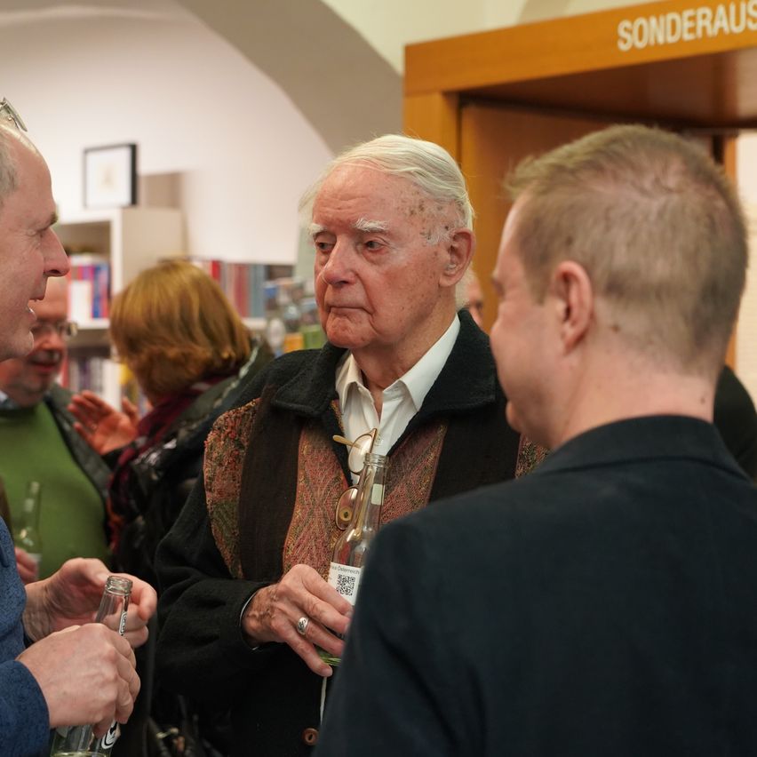 An older man with white hair and a patterned jacket stands in a library, holding a bottle and talking to two men in front of him.