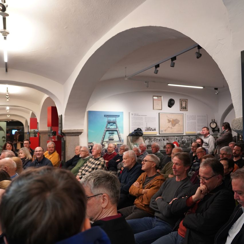 A room with arched ceilings and arched windows, where a large group of people are seated in rows, listening attentively. Some wear glasses, and others have their arms crossed.