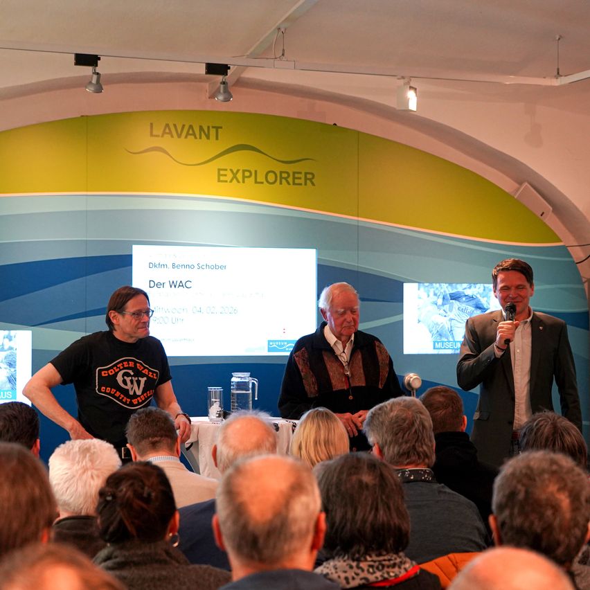 A man speaks into a microphone on a stage in front of a crowd. Behind him, a projector screen displays the words 'Der WAC'. Another man stands to his left, while another man sits to his right.