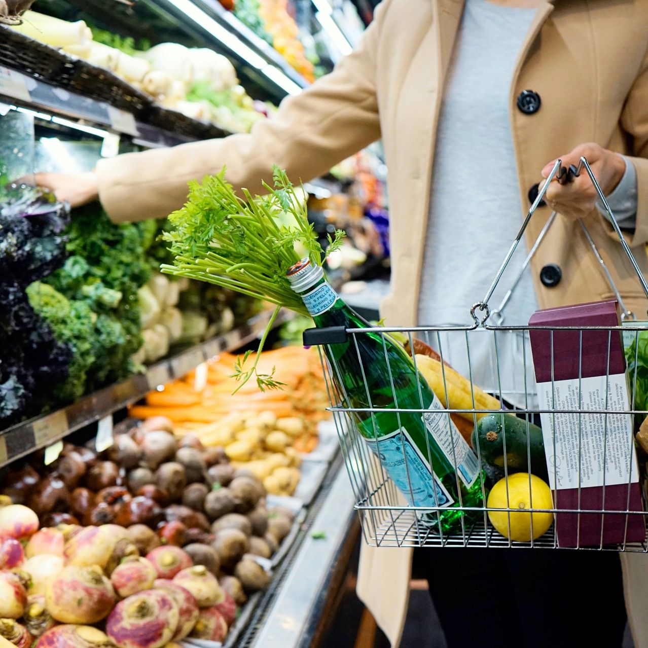 A woman in a supermarket, holding a shopping basket with vegetables and a bottle of wine.