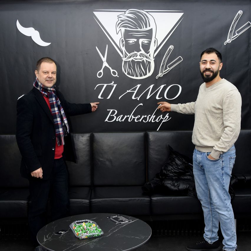 Two men stand in front of a black backdrop with a barber logo. The man on the left wears a scarf and a black coat. The man on the right wears a beige sweater and jeans.