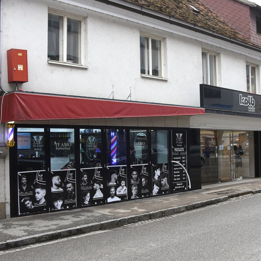 A white building with a red awning and a sign reading 'Kolb Fashion'. The shop has a glass door and black walls with pictures of men. A red box is attached to the wall.