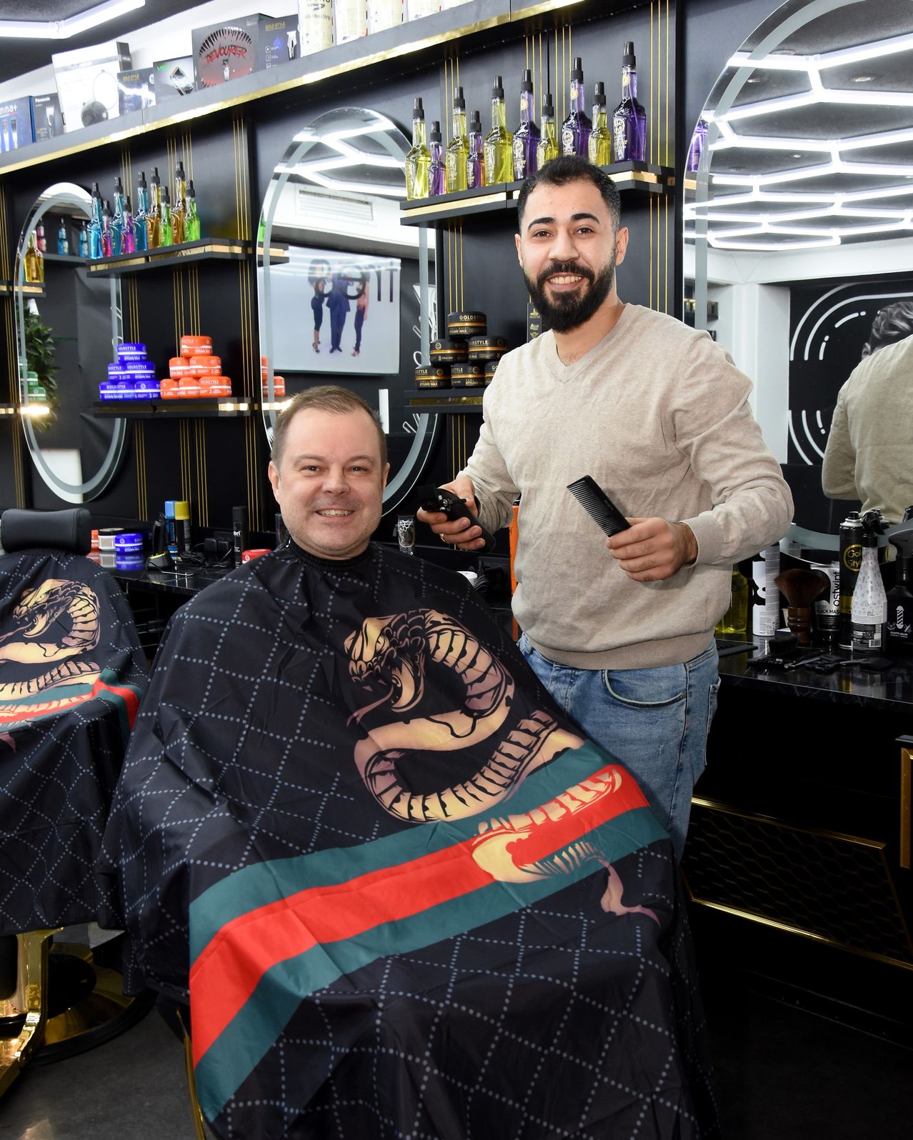 A man is sitting in a barber chair, wearing a cape with a snake design. A barber holds a comb and scissors. They are in a modern, well-lit barber shop with shelves filled with products.