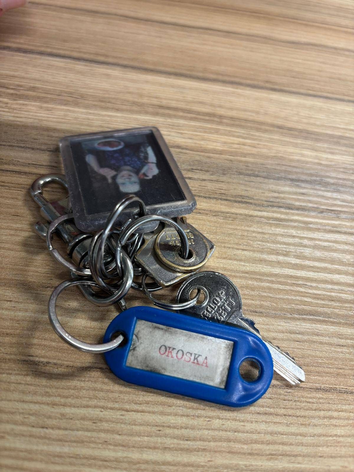A collection of keys with a photo frame attached, labeled 'OKOSKA', placed on a wooden surface.