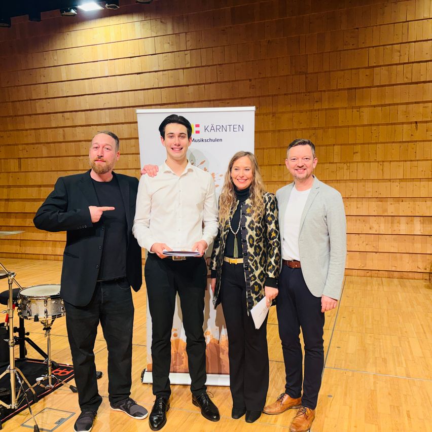 Vier Personen stehen nebeneinander in einem Musikraum. Ein Mann hält ein Zertifikat. Hinter ihnen ist ein Banner.
