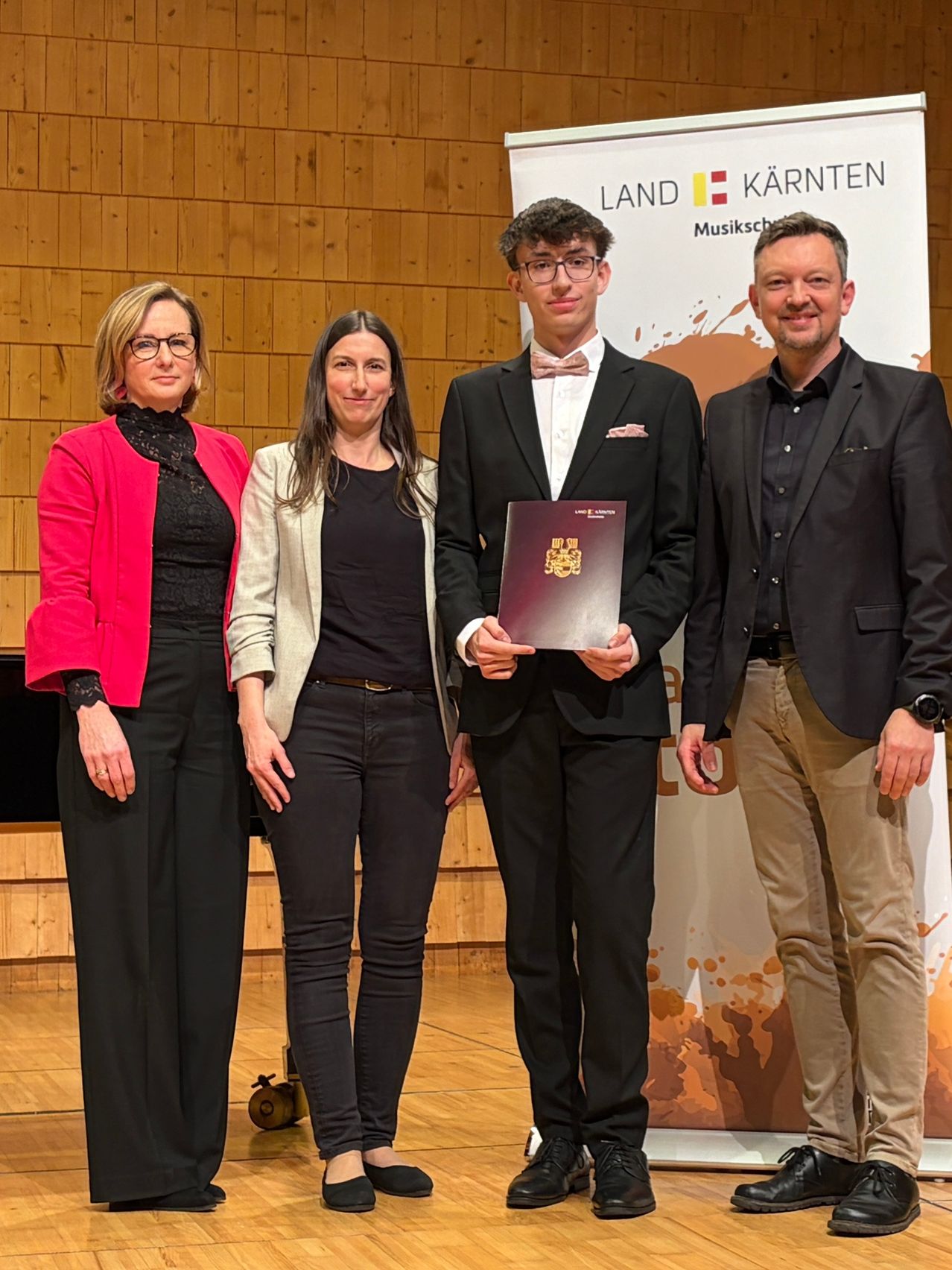 Vier Menschen stehen in einem Raum, wahrscheinlich bei einer Preisverleihung. Ein Mann in einem Anzug hält ein Zertifikat. Die Frauen links tragen eine Brille und einen Blazer. Hinter ihnen steht eine Holzwand mit einem Schild, auf dem 'LAND KARNTEN Musiksch' steht.