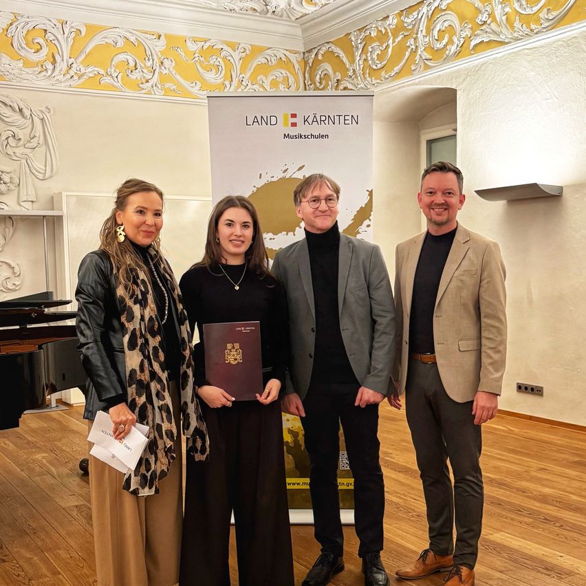 Drei Personen stehen in einem Raum und lächeln für ein Foto. Eine Frau hält ein Buch und Papier, eine andere ein Zertifikat. Ein Klavier und ein Banner mit 'LAND KARnten Musikschulen' sind im Hintergrund zu sehen.