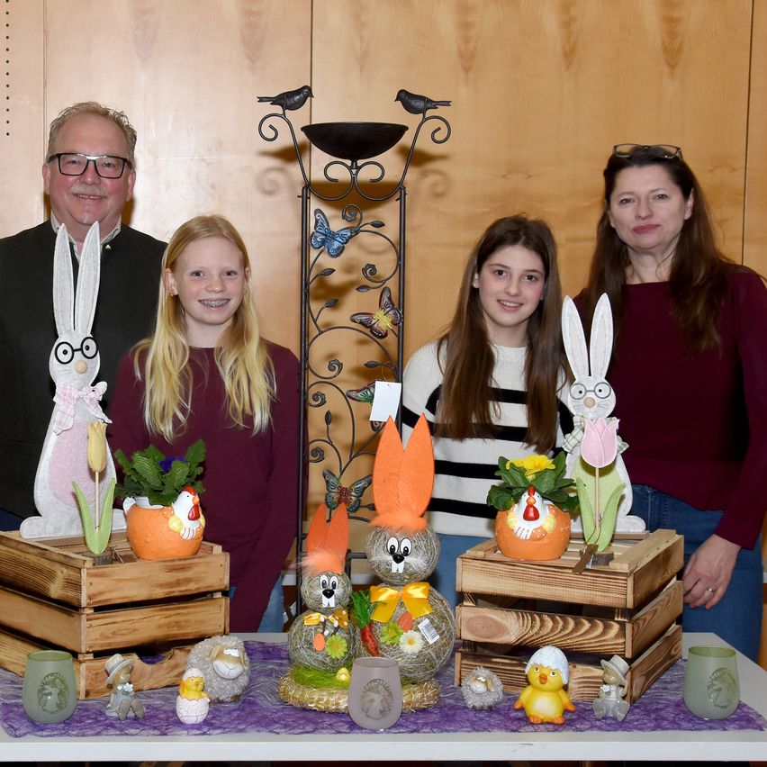 Eine Familie mit zwei Mädchen und einem Mann posiert hinter einem Tisch mit Osterdekorationen. Der Mann hat eine Hasenfigur auf seiner linken Seite und die beiden Mädchen haben Hasenfiguren auf ihrer rechten Seite. In der Mitte befindet sich eine große Hasendekoration mit orangefarbenen Ohren und einem gelben Band. Auf dem Tisch befinden sich auch zwei Holzkisten mit Pflanzen und anderen Osterdekorationen.