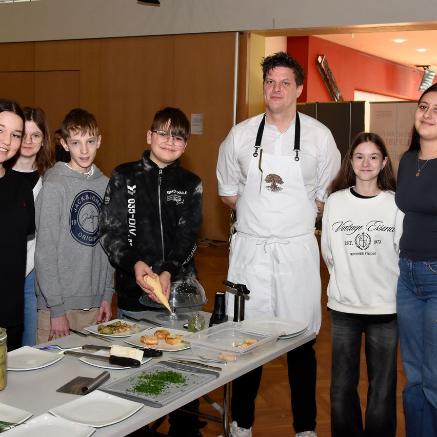 Eine Gruppe von Menschen, darunter vier Kinder, stehen um einen Tisch mit Essen und ein Koch in einer weißen Schürze. Sie lächeln und scheinen für ein Foto zu posieren.