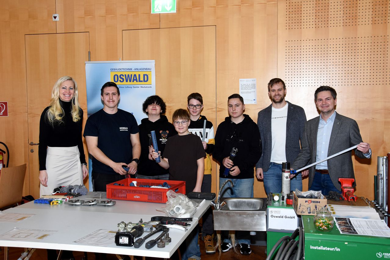 Eine Gruppe von Menschen steht um einen Tisch in einem Raum herum, mit verschiedenen Werkzeugen und Ausrüstung. Hinter ihnen befindet sich ein Banner mit dem Wort 'Oswald'. Es gibt zwei Waschbecken mit Wasserhähnen, und mehrere Kisten stehen auf dem Tisch. Ein Mann steht mit einem Werkzeug in der Hand.