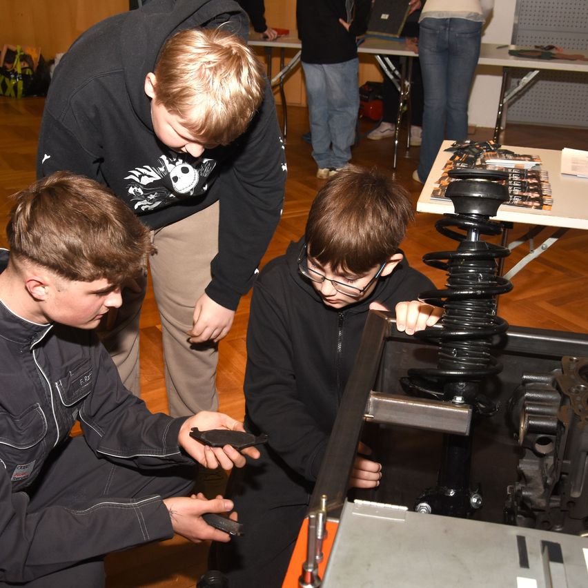 Drei junge Jungen arbeiten an einem mechanischen Gerät. Einer hält eine Bremsbelage, während ein anderer eine Schraubenfeder inspiziert. Der Junge in der Mitte betrachtet das Gerät genau. Hinter ihnen stehen andere Personen und Tische mit Gegenständen darauf.