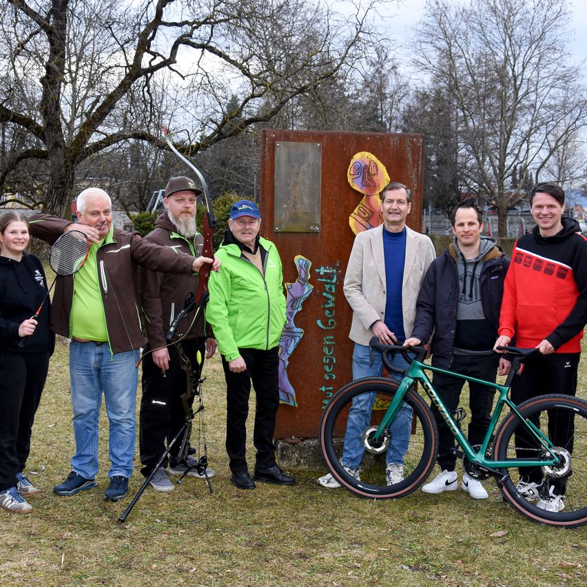 Sieben Personen stehen in einem Park. Eine Person hält ein Fahrrad. Eine andere Person hält einen Bogen mit Pfeil.