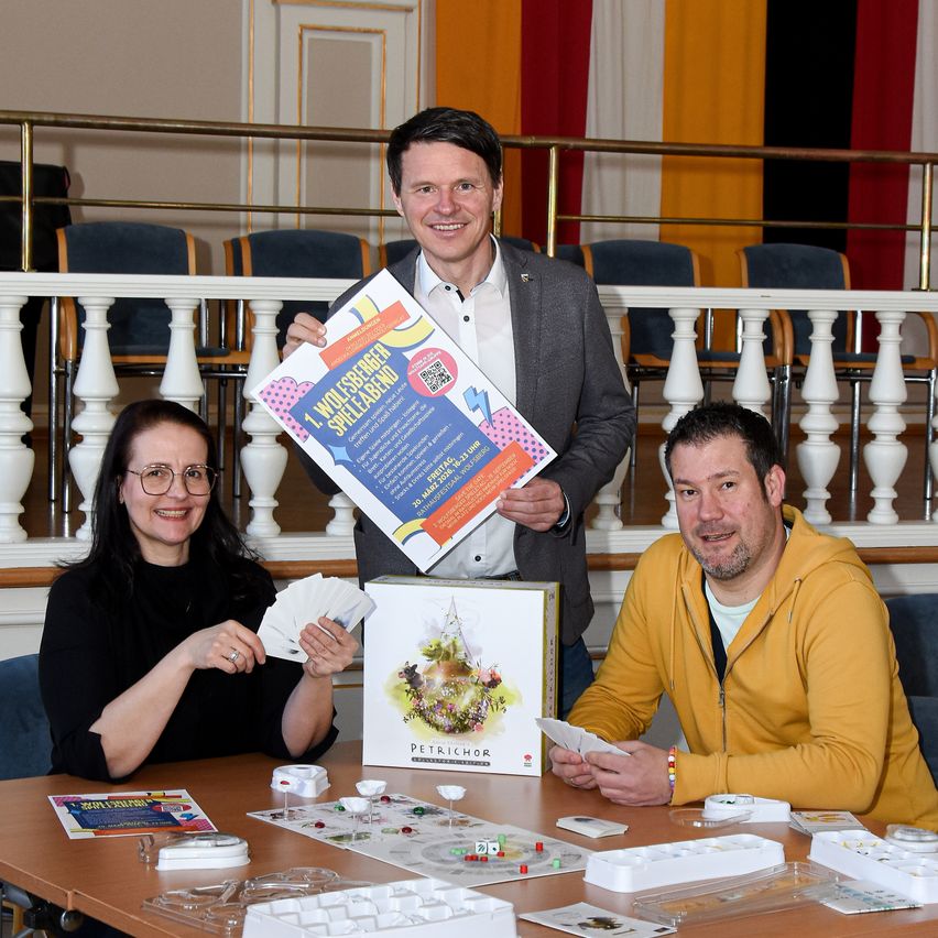 Drei Personen sitzen um einen Tisch herum und spielen ein Brettspiel. Ein Mann, der ein Poster hält, steht hinter ihnen und lächelt. Die Spielebox ist mit 'Petrichor' beschriftet. Der Tisch ist mit verschiedenen Spielkomponenten bedeckt.