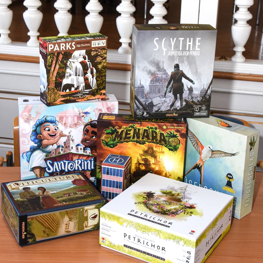 Eine Sammlung von Brettspielen, darunter Scythe, Parks, Santorini, Viticulture und Petrichor, auf einem Holztisch ausgestellt.