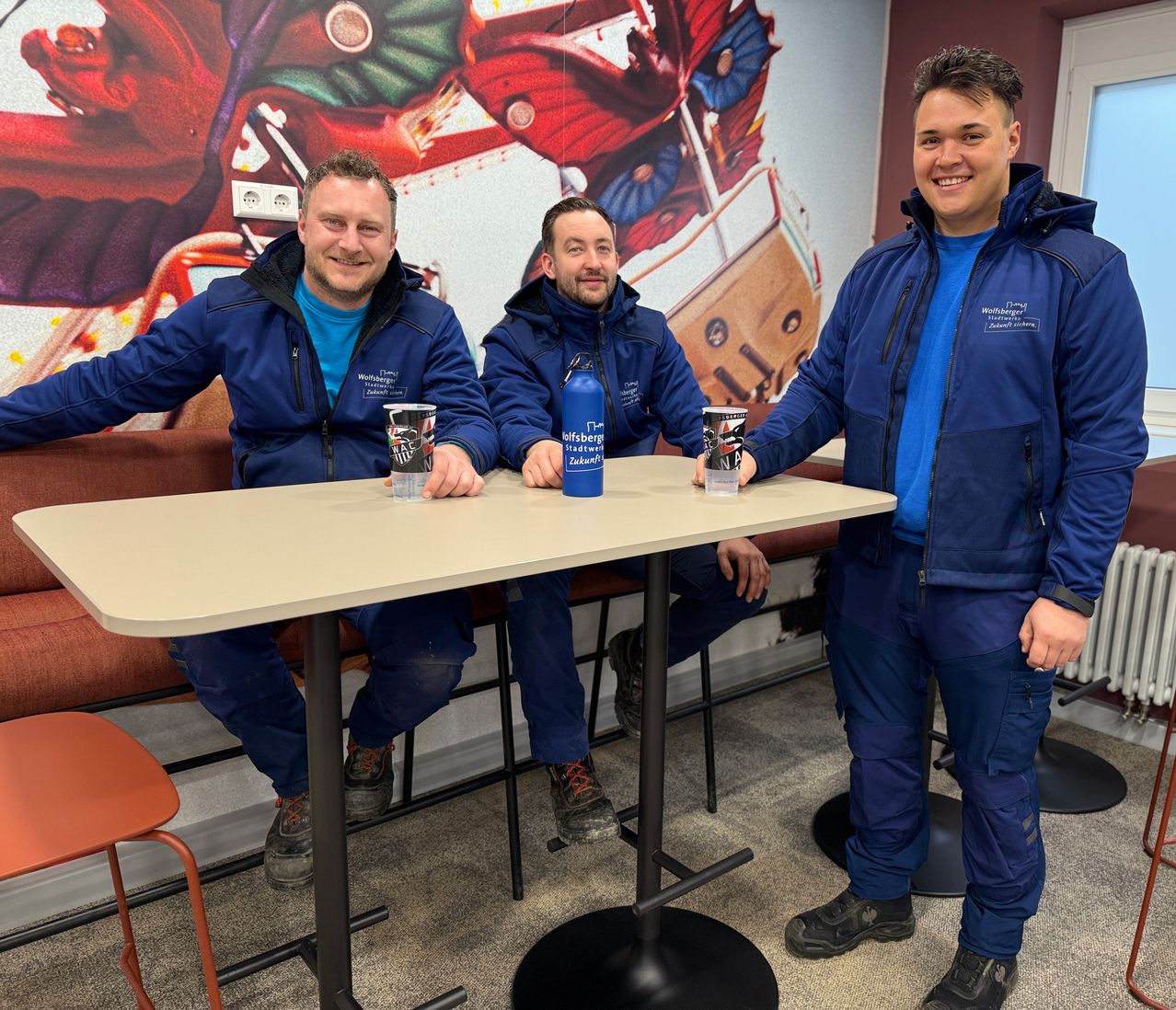 Drei Männer in blauen Jacken sitzen lächelnd an einem Tisch. Eine blaue Wasserflasche steht in der Mitte, und sie haben Tassen vor sich. Die Wand hinter ihnen hat ein großes Gemälde.