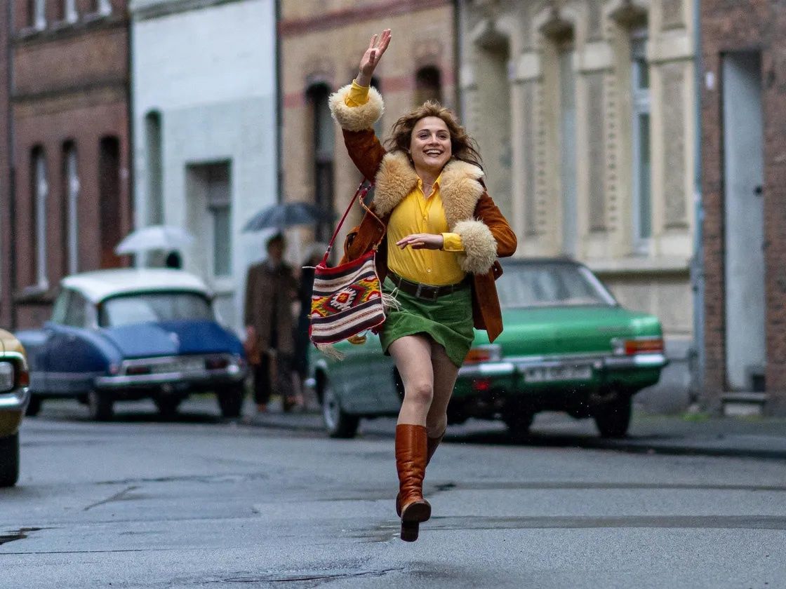 Eine Frau läuft fröhlich auf der Straße, die Hand gehoben, trägt einen Pelzmantel, ein gelbes Hemd, einen grünen Rock und braune Stiefel. Hinter ihr gehen zwei Personen mit Regenschirmen, und zwei Autos sind geparkt.