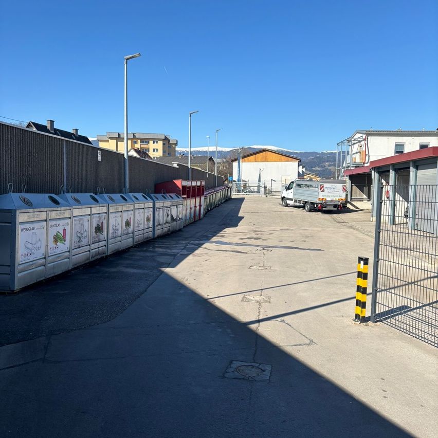 Ein Parkplatz mit Mülleimern und einem weißen LKW, der vor einem Gebäude mit einem Berg im Hintergrund geparkt ist.