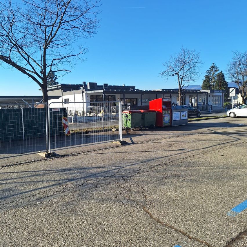 Ein leerer Parkplatz mit Bäumen und einem Gebäude im Hintergrund. Ein roter Mülleimer steht neben einem Zaun. Ein weißes Auto ist rechts geparkt.