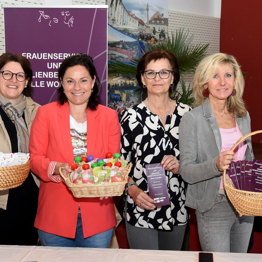 Vier Frauen stehen lächelnd vor einem Tisch und halten Körbe mit Produkten. Hinter ihnen steht ein violetter Banner mit der Aufschrift 'Frauenservice und familienfreundliche Wolfsberg'.