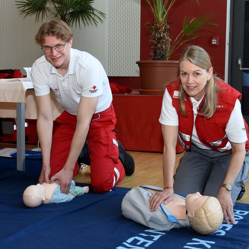 Zwei Personen in roten Westen demonstrieren Herz-Lungen-Wiederbelebung an einem Mannequin, eine kniet und die andere steht. Sie sind drinnen, mit einer roten Wand, Topfpflanzen und einem Tisch mit einem Tuch im Hintergrund.