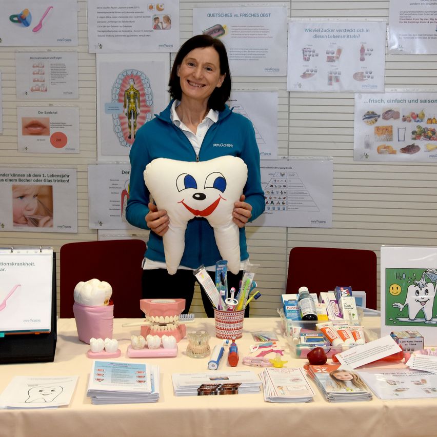 Eine Frau steht hinter einem Tisch mit Zahnartikeln, lächelt und hält einen großen Zahn-Plüsch. Der Tisch ist mit einer weißen Tischdecke bedeckt und zeigt verschiedene Zahnpflegeprodukte.