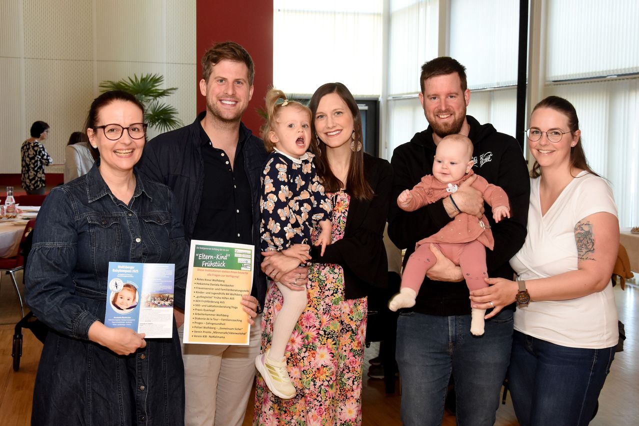 Eine Familie posiert mit einem Zertifikat und Broschüren. Die Familie besteht aus zwei Erwachsenen, zwei Kindern und einem Baby. Sie lächeln und scheinen bei einer Preisverleihung zu sein.