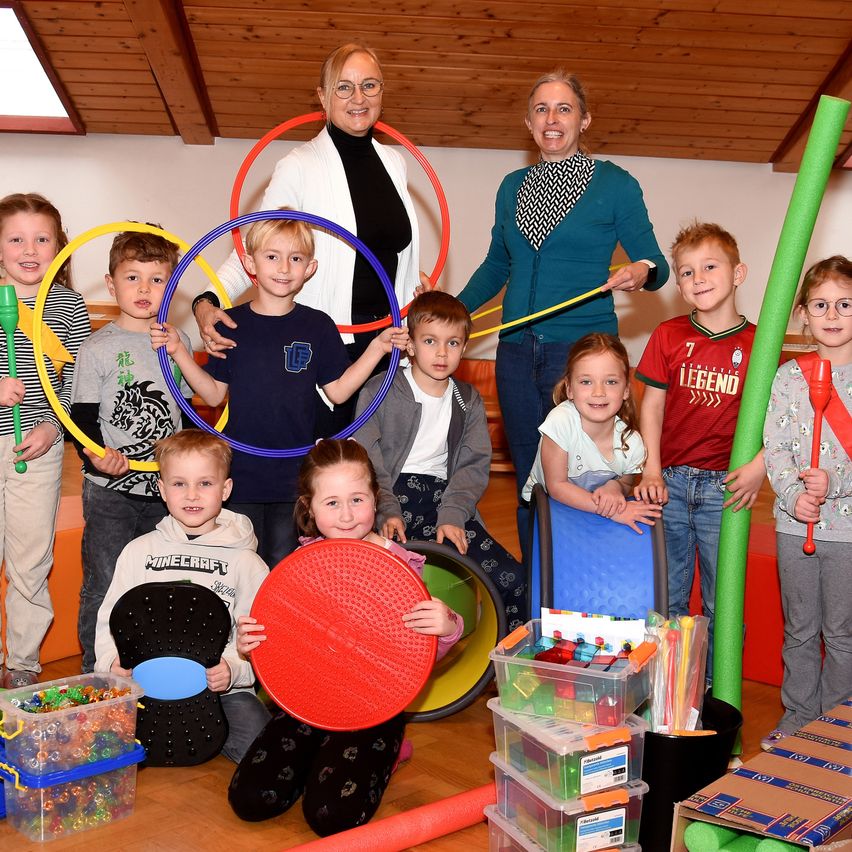 Eine Gruppe von Kindern und zwei Erwachsenen sind in einem Spielzimmer. Die Kinder halten verschiedene Spielzeuge, darunter Hula-Hoops und eine rote Scheibe. Die Erwachsenen lächeln und scheinen für ein Foto zu posieren.