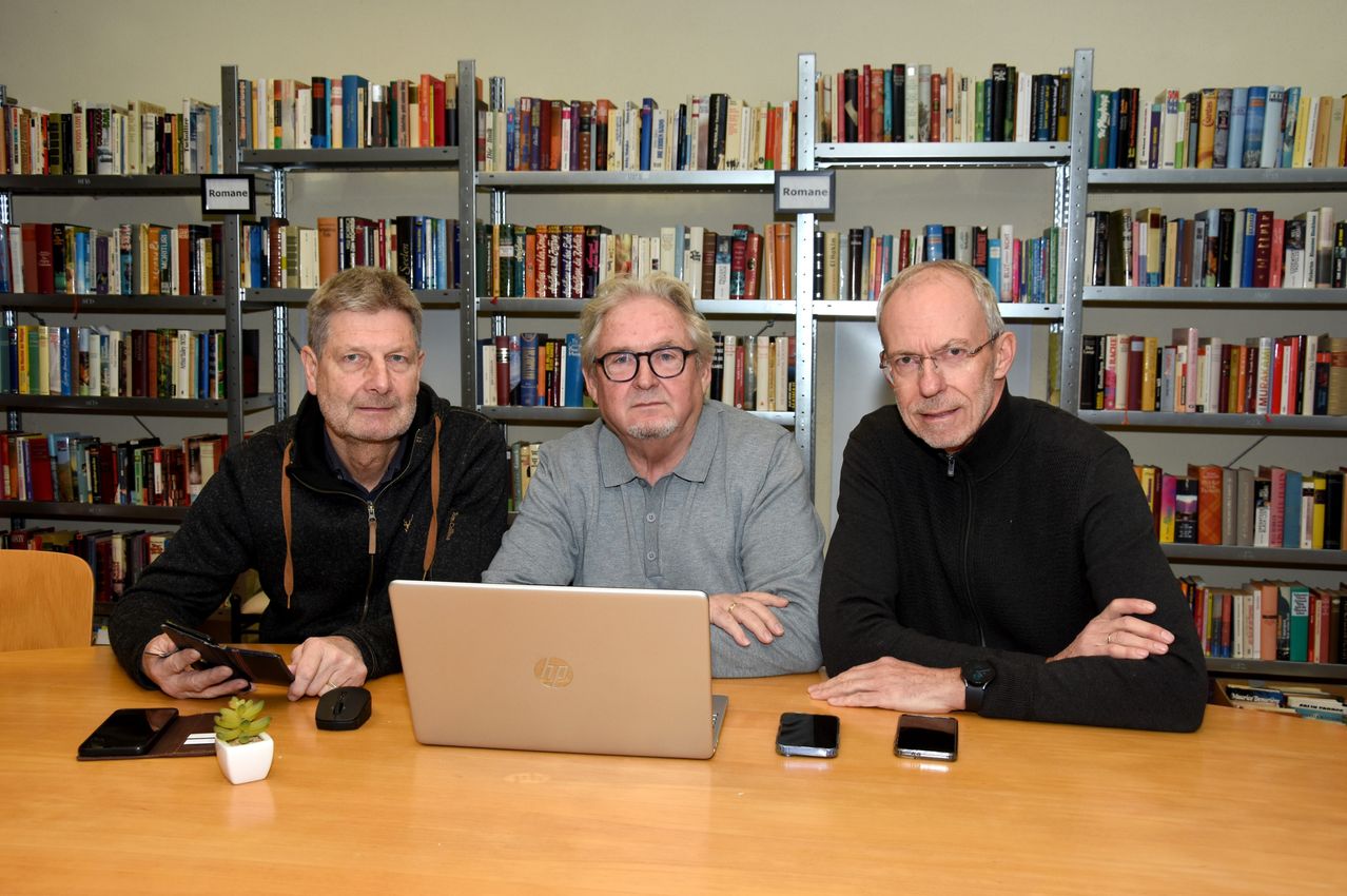 Three older men are sitting in front of a desk with a laptop, phones, and a small plant. They are in a library with shelves full of books behind them.
